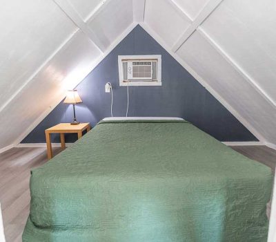 A small attic bedroom with white sloped ceilings, a double bed covered in a green quilt, a wooden nightstand with a lamp, and a wall-mounted air conditioner above the bed on a blue accent wall.
