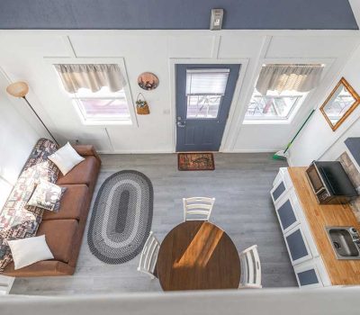 A cozy, bright tiny house interior seen from above, featuring a brown couch with white pillows, a round dining table with four chairs, a small kitchen area, and light gray wood floors. Sunlight streams through the windows.