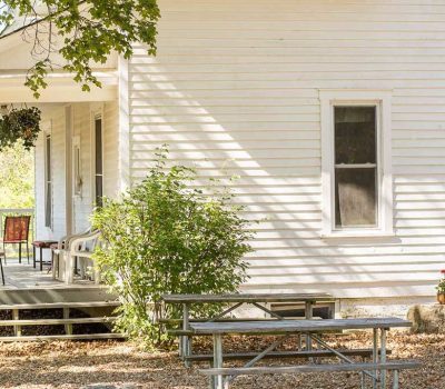 A white wooden house with a porch, red and white chairs, hanging plants, and a picnic table in the yard. Sunlight casts shadows across the siding and ground, and leafy branches hang above.