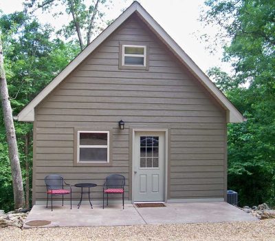 A small beige cabin with a peaked roof sits in a wooded area. The front has a glass door, two windows, a wall lamp, and a patio with two black chairs and a round table.