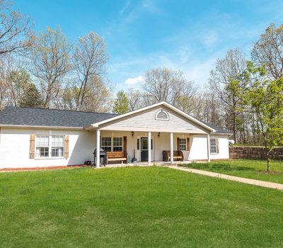 A single-story white house with a dark roof sits on a grassy lawn, surrounded by trees with spring foliage under a blue sky. A small porch and walkway lead to the front door.
