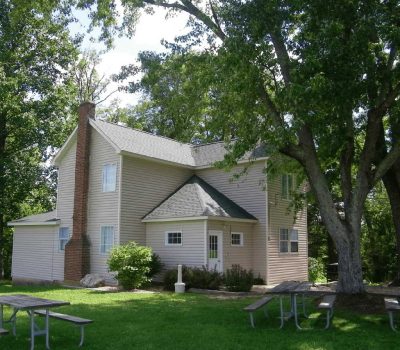 A two-story beige house with a brick chimney sits among tall trees and green grass, with picnic tables in the yard under the shade.