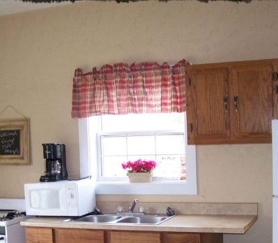 A small kitchen with a sink, microwave, coffee maker, stove, and refrigerator. A window with a red plaid curtain and a potted plant is above the sink. Wooden cabinets are on the wall. A chalkboard hangs to the left.