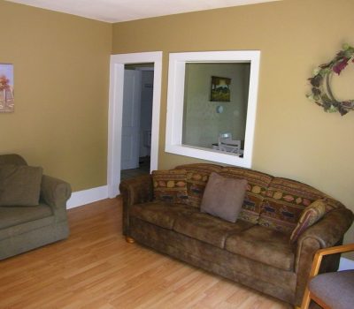 A cozy living room with light wood flooring, a brown patterned sofa, an armchair, and a loveseat. The walls are tan with white trim, and there’s a wall wreath, a framed painting, and a window to another room.