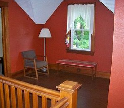 A small room with red walls, a wooden floor, and a window with white curtains. There is a simple gray chair with wooden arms, a striped bench, a floor lamp, and a wooden stair railing in the foreground.